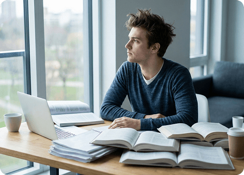 צעיר יושב ליד שולחן עם ספרים ומחשב נייד ומביט דרך החלון